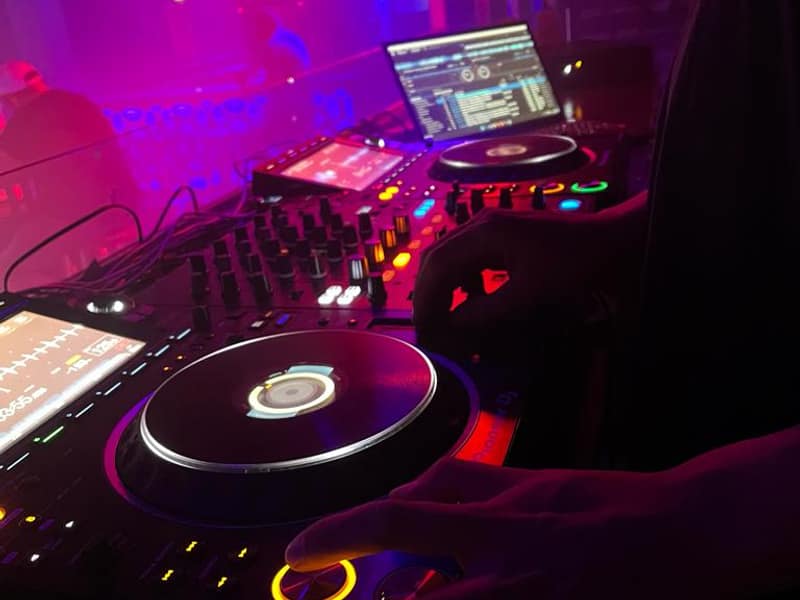 Nahaufnahme eines DJ-Pults mit CDJ, Mixer und Laptop in pinkfarbenem und blauem Bühnenlicht
