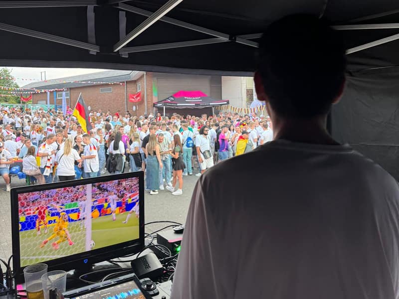 DJ-Pult unter einem Pavillon mit Public-Viewing-Bildschirm und großer Menschenmenge im Hintergrund
